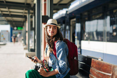 Happy woman wearing hat sitting on bench at tram station