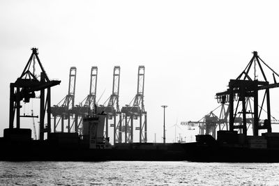 Pier and cranes at harbor against sky in black and white