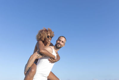 Low angle view of man piggybacking cheerful girlfriend against clear blue sky