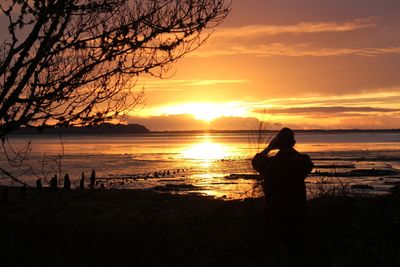 Silhouette woman photographing sea against sunset sky