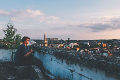 Man sitting by river against sky during sunset