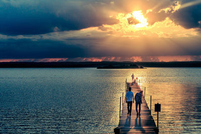 People on pier over river against cloudy sky at sunset