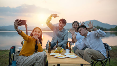 Cheerful friends taking selfie while sitting at lakshore