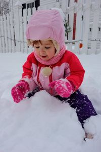 Girl in warm clothing playing while sitting on snow covered field against fence