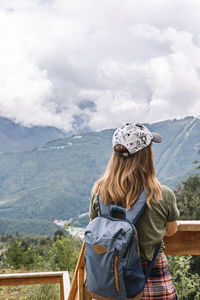 Rear view of young blonde woman in cap looking at view of mountains on wooden viewpoint, hiking 