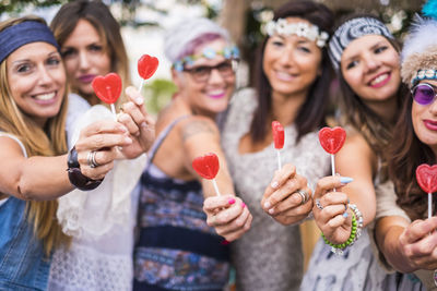 Portrait of happy friends holding heart shape lollipops while standing in yard