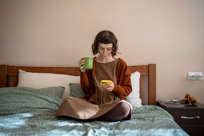 Pensive teenager sitting on bed with cup of tea, reading news in internet, hanging in social network