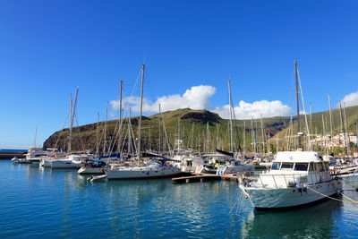 Sailboats moored in sea by mountain against blue sky