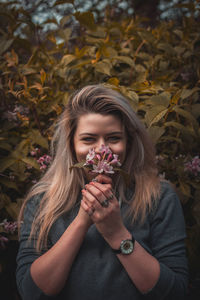 Portrait of smiling young woman holding flowers against tree 