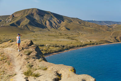 Young, happy woman stands on rock with her arms raised and looks at sea below.