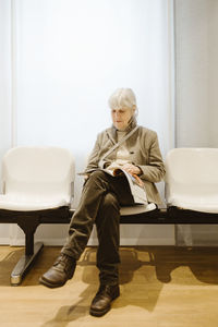 Full length of senior woman reading magazine while sitting on chair in waiting room at hospital