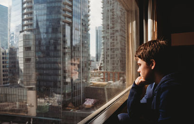 Tween boy looking out a window at tall buildings of the city outside.