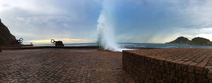 Waves splashing at shore against cloudy sky