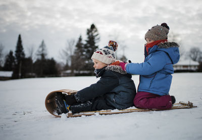 Side view of siblings on sled against cloudy sky