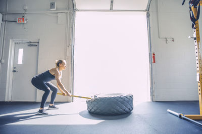 Side view of woman hammering tire while exercising in brightly lit gym