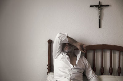 Adult man in white shirt in room lying on bed with cross on wall