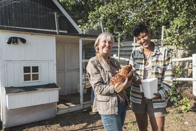 Portrait of smiling multiracial mother and daughter with chicken at farm on sunny day