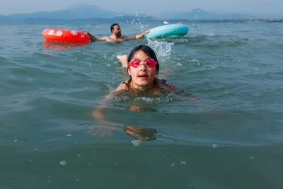 Girl in pink goggles swimming in the lake with father playing with another sibling behind