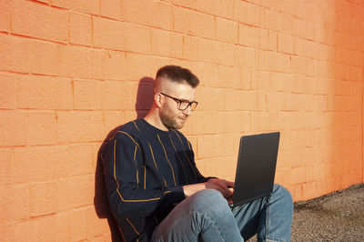 Man using laptop while sitting against brick wall
