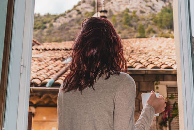 Rear view of woman holding coffee cup standing by window