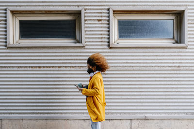 Young woman with protective face mask using mobile phone by wall during covid-19