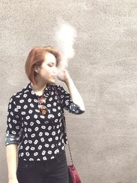 Beautiful young woman smoking while standing against wall