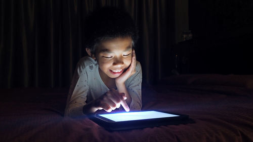 Smiling boy using digital tablet while lying on bed in darkroom