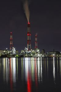 Smoke stacks in illuminated factory by lake against sky at night