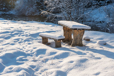 Seats on snow covered landscape by trees and water during winter