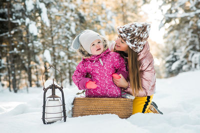 Mother embracing with daughter while sitting on snow covered land during winter