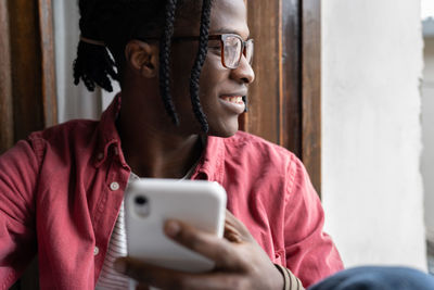 Positive satisfied african american man with smartphone sits near window coming up with sms