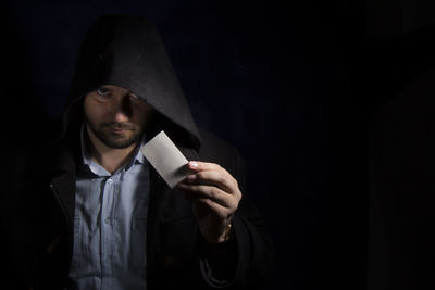 Portrait of serious man wearing hooded shirt while holding blank business card against black background