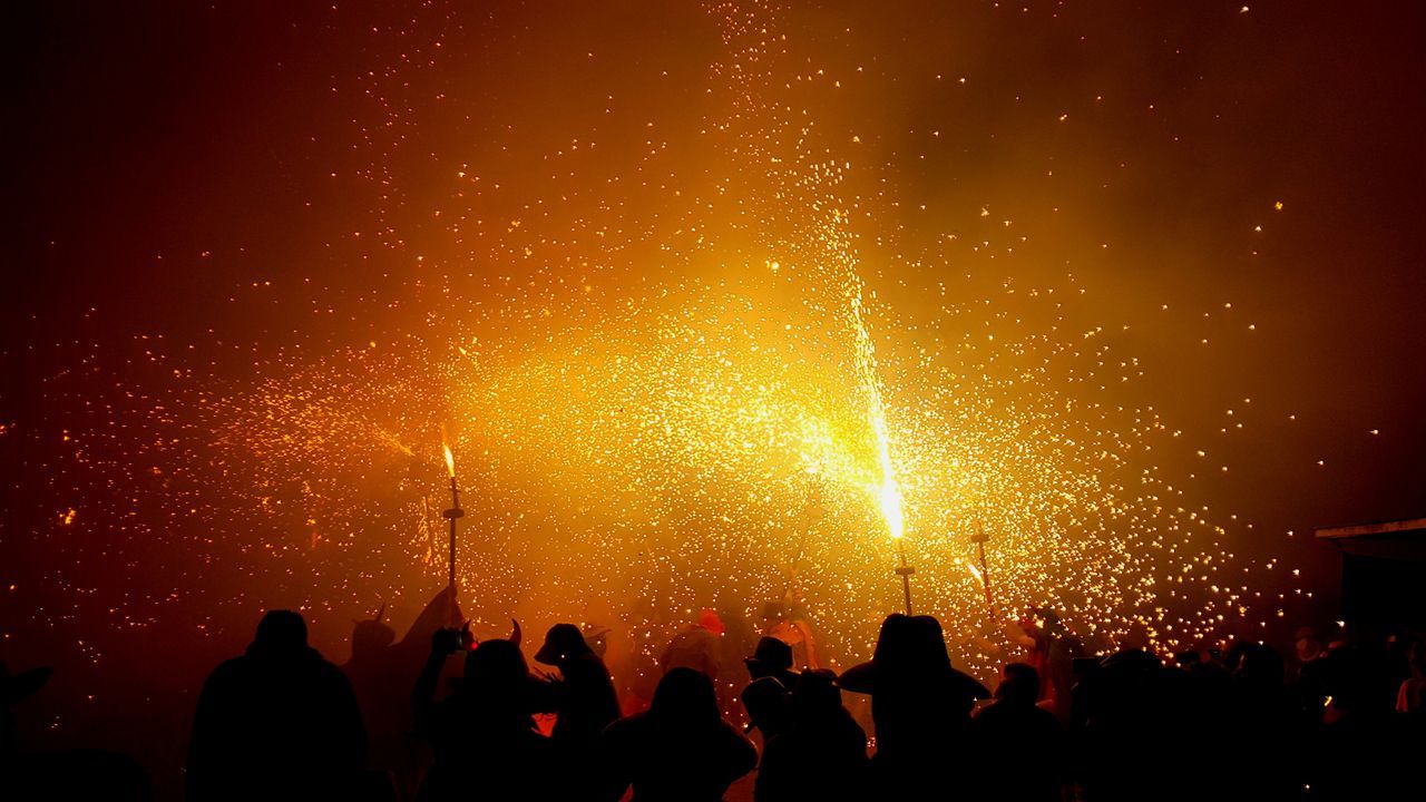 exploding, large group of people, arts culture and entertainment, night, event, glowing, celebration, firework display, real people, orange color, smoke - physical structure, firework - man made object, silhouette, burning, men, leisure activity, outdoors, women, crowd, low angle view, lifestyles, illuminated, firework, sky, people