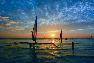 Silhouette outrigger sailboats in sea at beach against sky during sunset