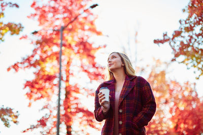 Young woman holding coffee while standing against autumn trees