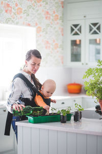 Side view of female blogger watering plants while carrying daughter in kitchen at home
