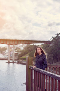 Young woman standing by railing on bridge against sky