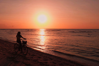 Man riding bicycle on sea against sky during sunset