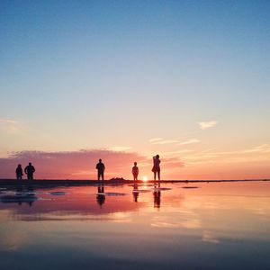 People standing with reflection on wet shore at beach against sunset sky