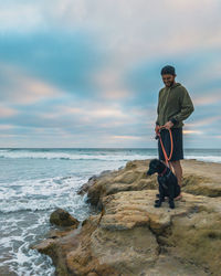 Man with dog standing on rock at beach against cloudy sky