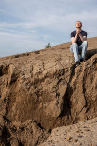 Young man sitting on rock against sky