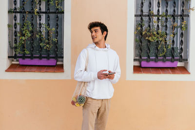 Young male standing on city street with eco friendly mesh bag with groceries and using smartphone while looking away