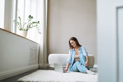 Young woman in jeans working on laptop sitting on carpet at home, view from doors