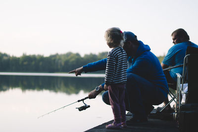 Side view of daughter and father fishing in lake against sky