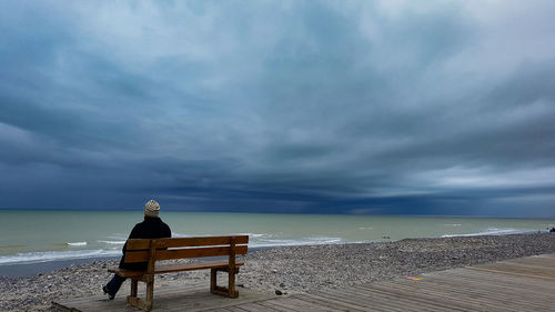 Rear view of man sitting at bench at beach against sky