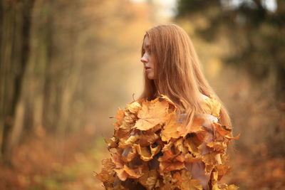 Young woman covered with leaves standing at forest during autumn