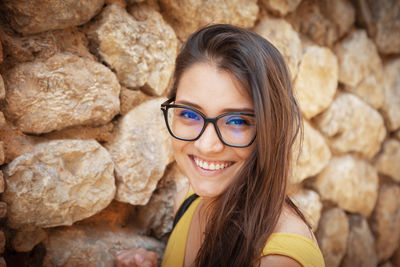 Young woman standing in front of a stone wall. wearing glasses smiling at the camera