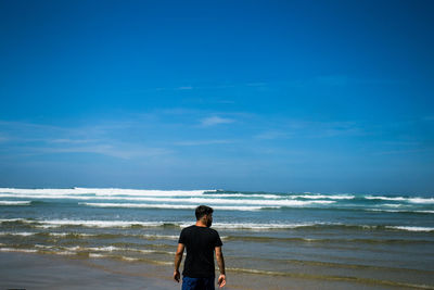 Rear view of mid adult man standing at beach against blue sky