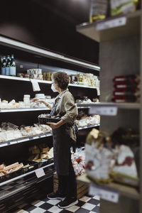 Female owner arranging food product on rack at deli shop during pandemic