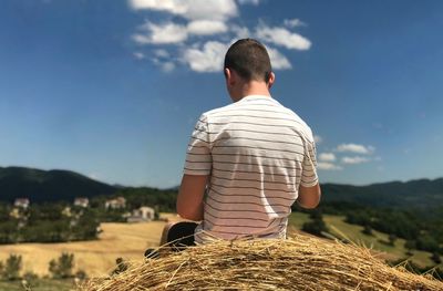 Rear view of man sitting on hay bale against sky during sunny day